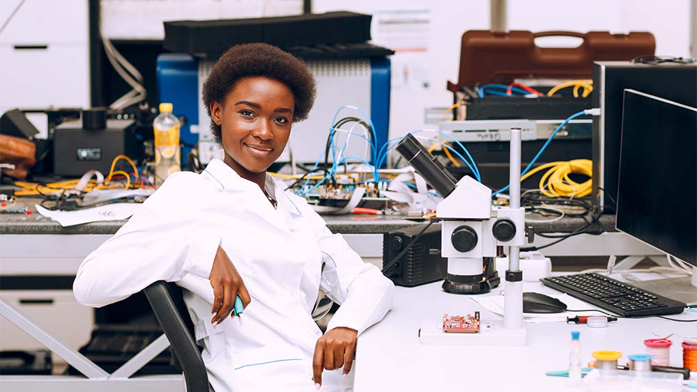 Une personne portant un manteau blanc regarde la caméra. Un ordinateur et un microscope se trouvent devant lui. De nombreux gadgets électroniques et câbles de différentes couleurs sont visibles en arrière-plan. 