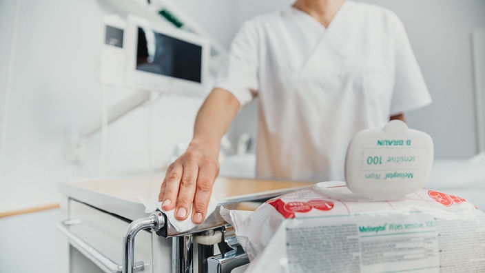 Infirmière debout à une table, face à un petit garçon souriant. Sur la table, une petite ambulance mannequin est poussée par la femme, surveillée par le garçon. Au premier plan, un paquet de lingettes Meliseptol.