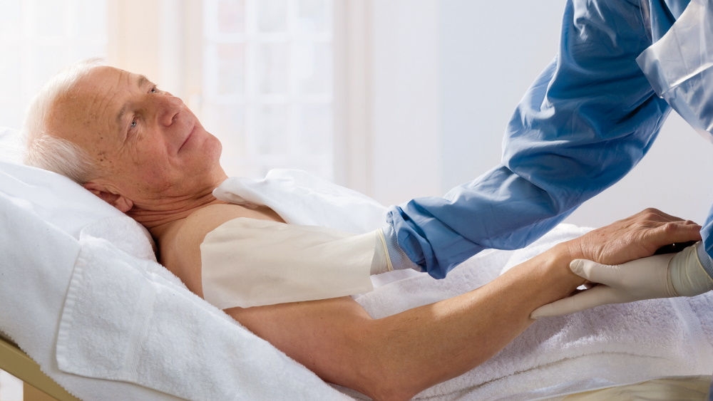 Patient dans un lit d’hôpital. L’infirmière décolonise le haut du bras du patient à l’aide d’un chiffon.