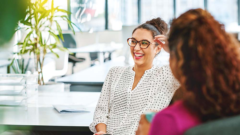 À droite, une femme est assise avec le dos de sa tête, à gauche, une femme est assise devant elle et regarde de côté en riant. En arrière-plan, un bureau