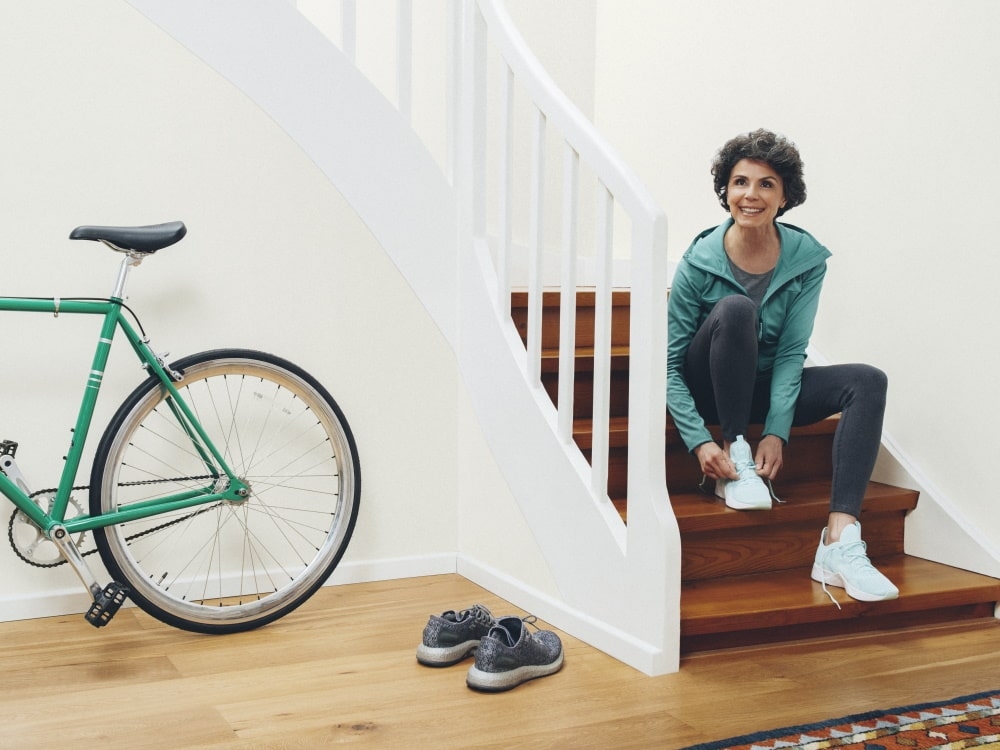 Rechts sitzt eine Frau auf der Treppe und bindet ihre Schuhe, links ein Fahrrad und ein weiteres Paar Schuhe