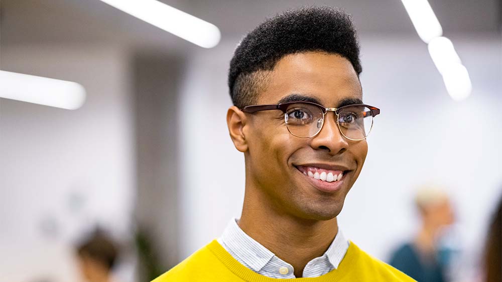 Gros plan sur une personne souriante avec un haut jaune et des lunettes debout dans un environnement lumineux. 