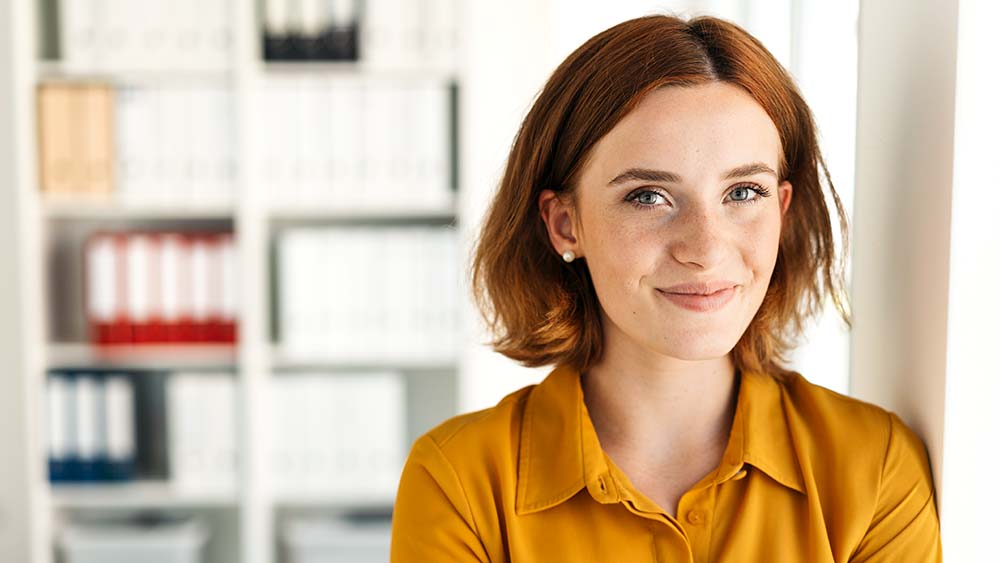 Une femme souriant en toute confiance, regardant directement la caméra, des étagères à livres sont visibles en arrière-plan. 